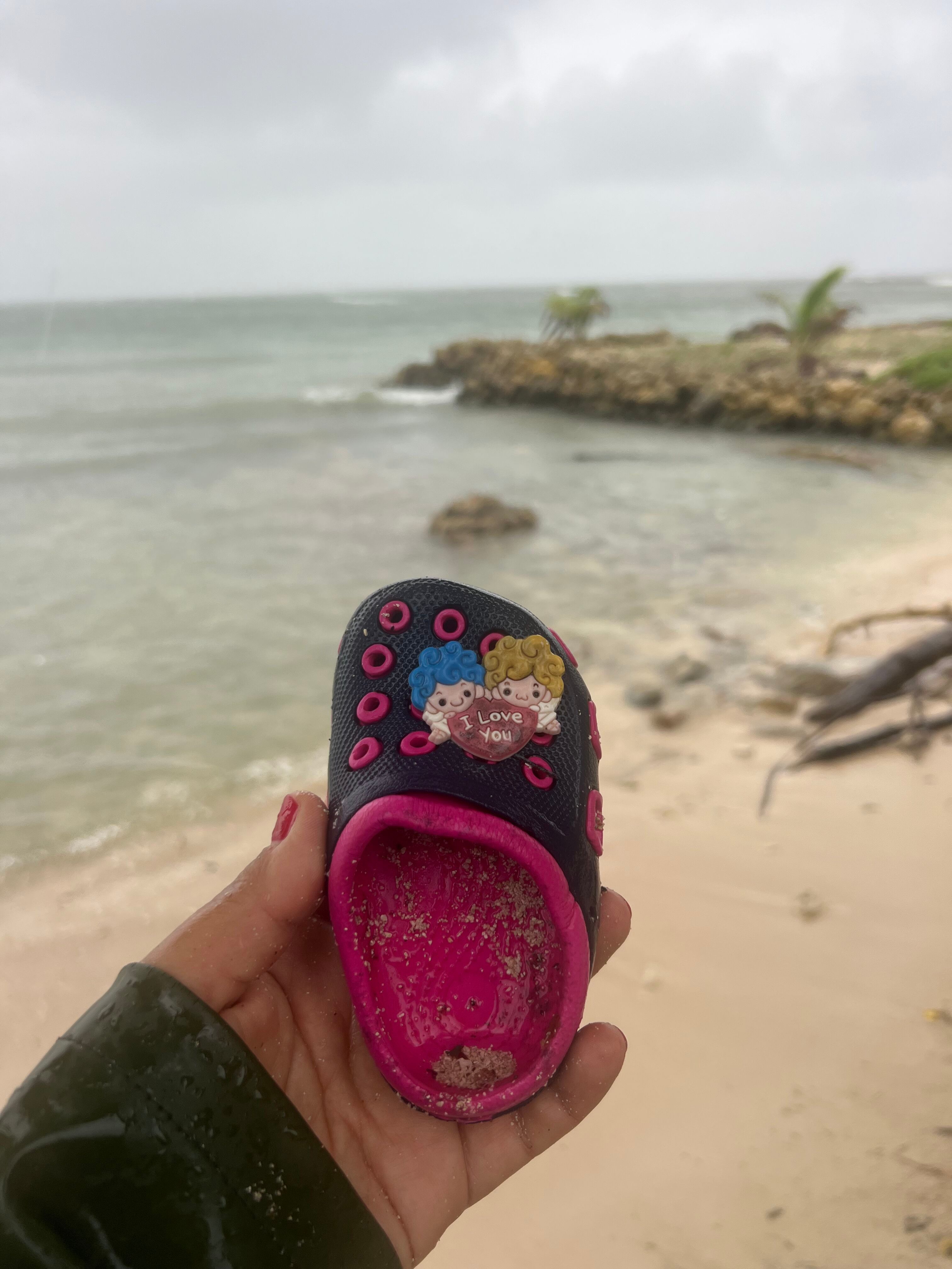 A small pink and black child's clog shoe found on a beach, featuring a cartoon 'I Love You' charm.